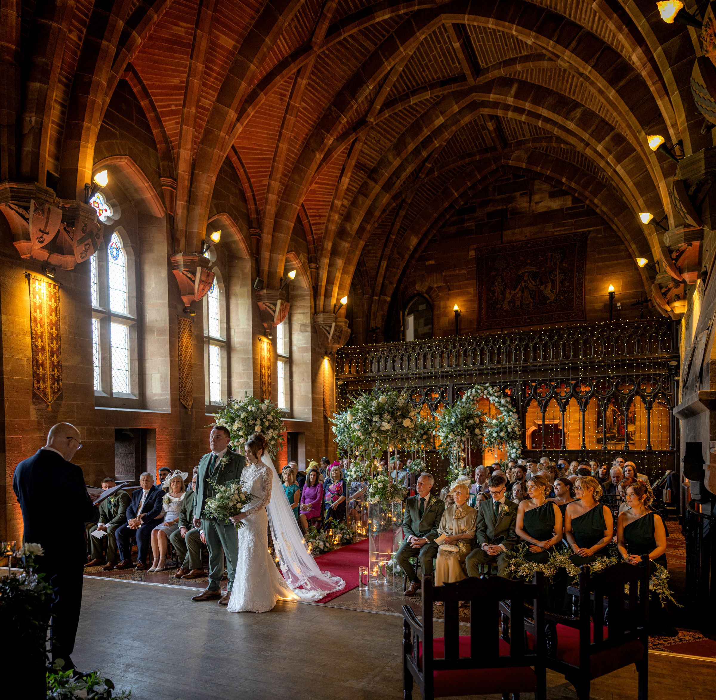 Bride in a white gown and groom exchange vows at a wedding ceremony in a historic church with ornate timber arches and floral arrangements.