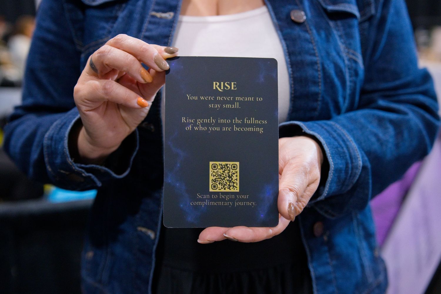 Woman in a denim jacket holds a dark navy affirmation card reading Rise with a QR code at the bottom.
