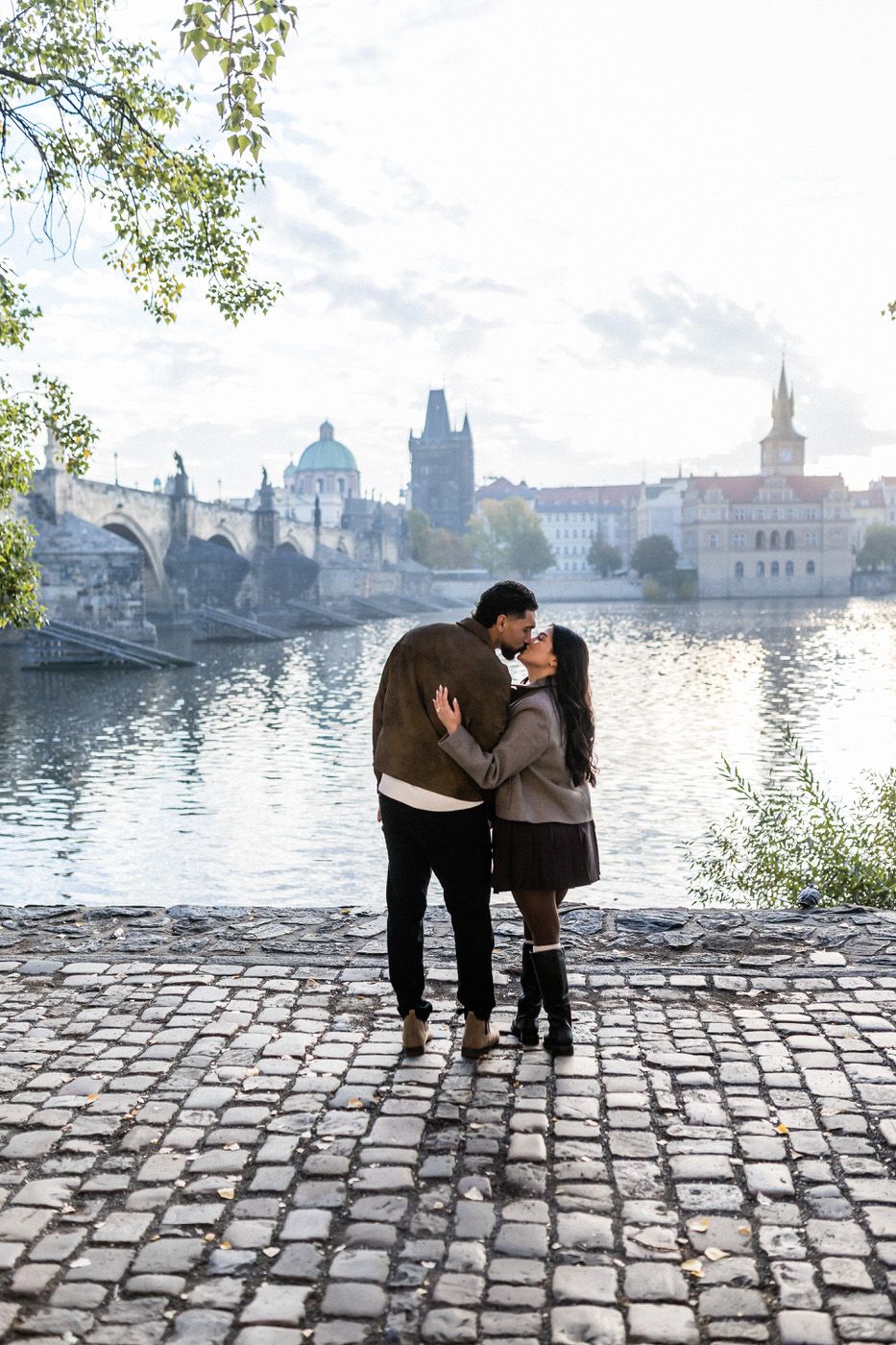 Couple kissing and embracing on a cobblestone riverbank in Prague with historic bridge towers and spires across the Vltava River.
