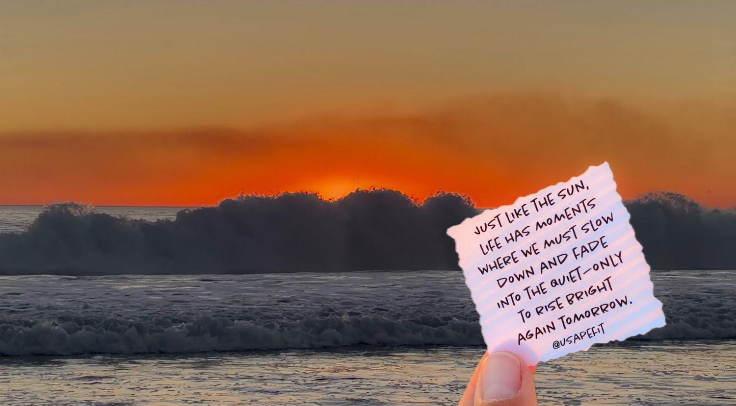Hand holding an inspirational note card against a stunning orange and pink ocean sunset with crashing waves.