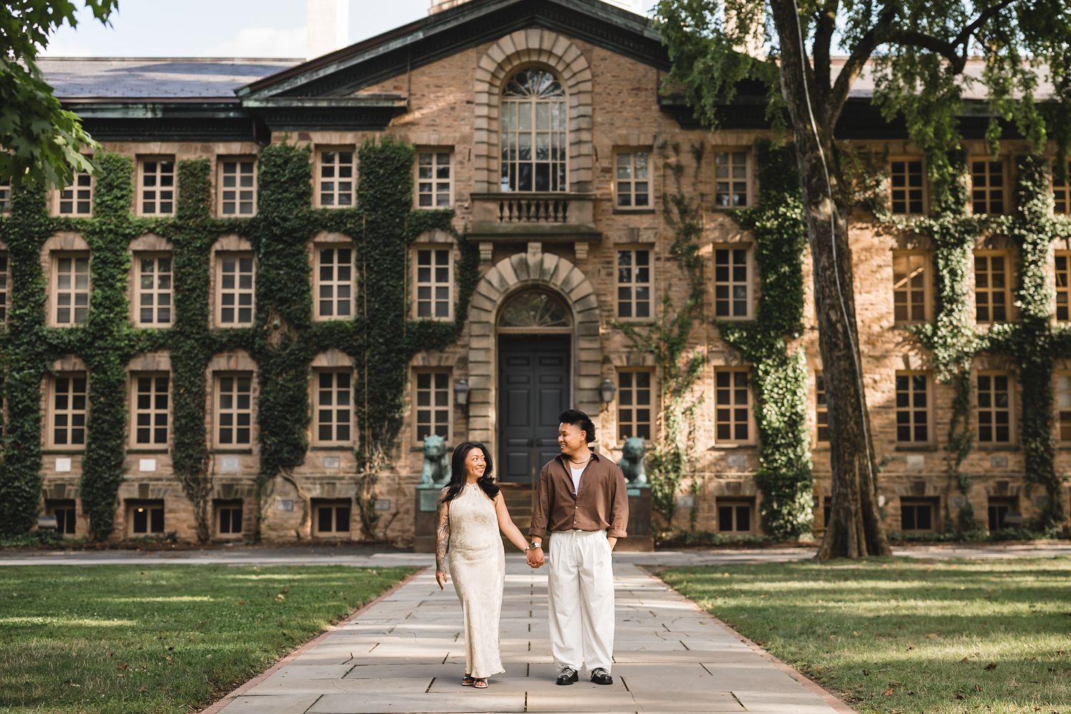 Couple holding hands walking on a pathway in front of an ivy-covered brick building at Princeton University.