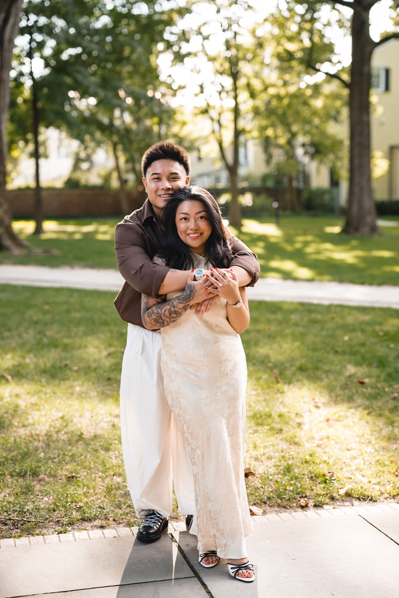 A couple embraces outdoors, the woman in a white lace dress and the man in a brown jacket, surrounded by green trees.