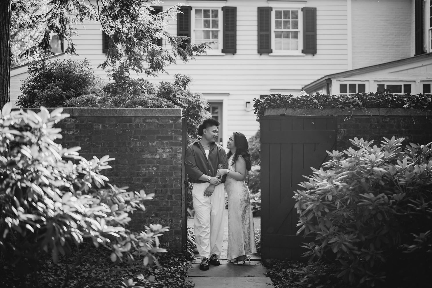 A couple shares a romantic moment at a garden gate surrounded by lush greenery near a classic white home.