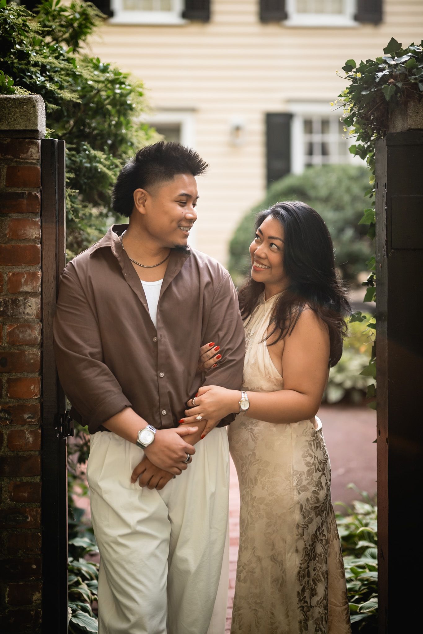 A couple holds hands and smiles at each other standing between brick pillars and lush greenery outside a home.