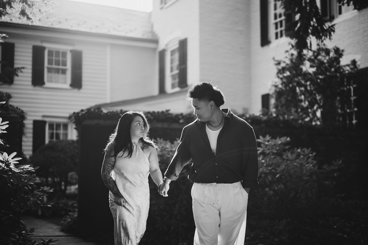 A couple holds hands walking through a garden path near a white colonial home in a romantic black and white photo.
