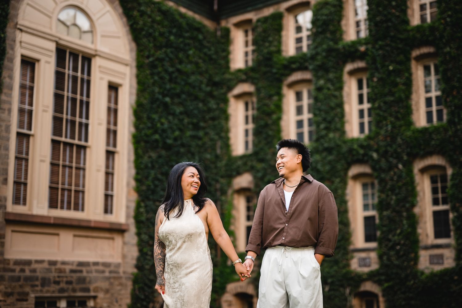 Couple holding hands smiling before ivy-covered stone building, elegant engagement photo.