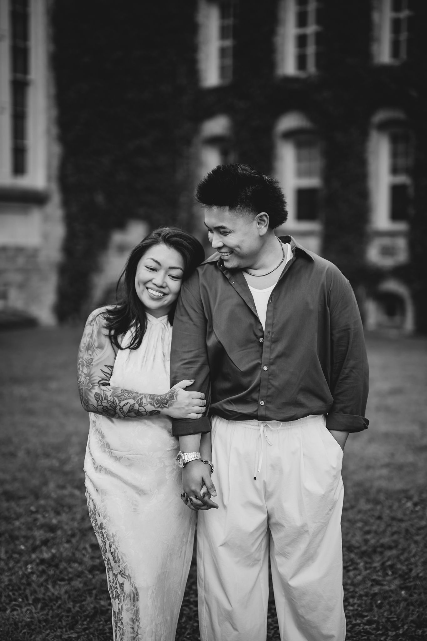 A black and white photo of a smiling couple embracing on a lawn in front of an ivy-covered building.