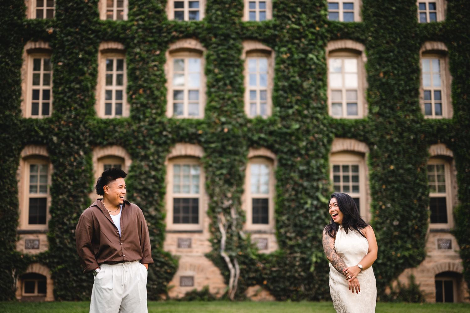 Couple standing apart smiling at each other on a green lawn before an ivy-covered stone building.