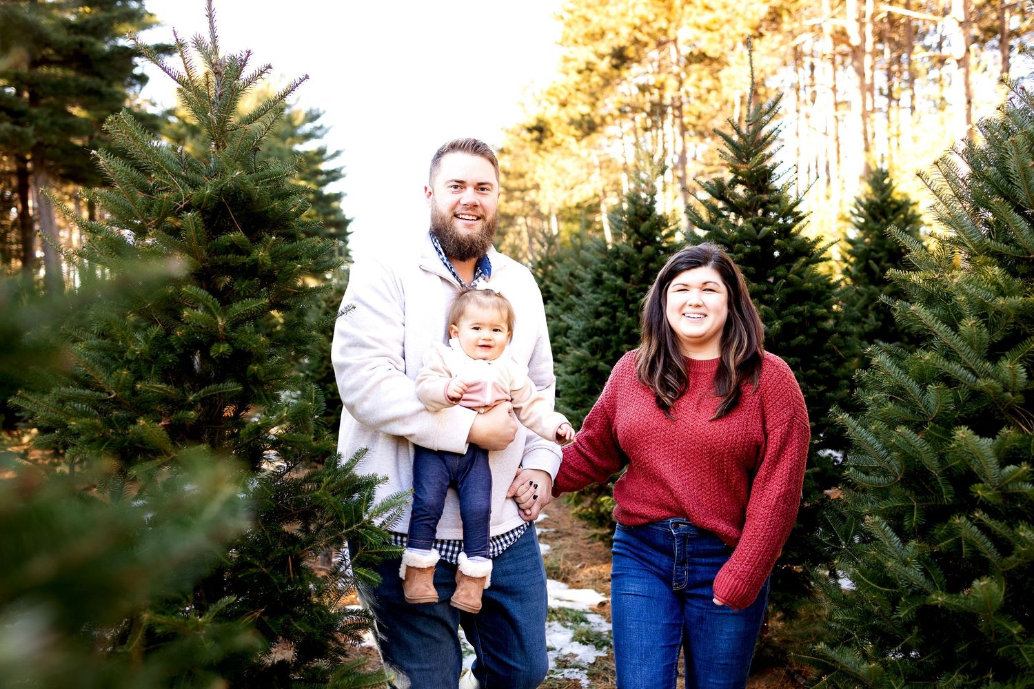 Family of three smiling among Christmas trees at a tree farm, parents holding baby girl outdoors.