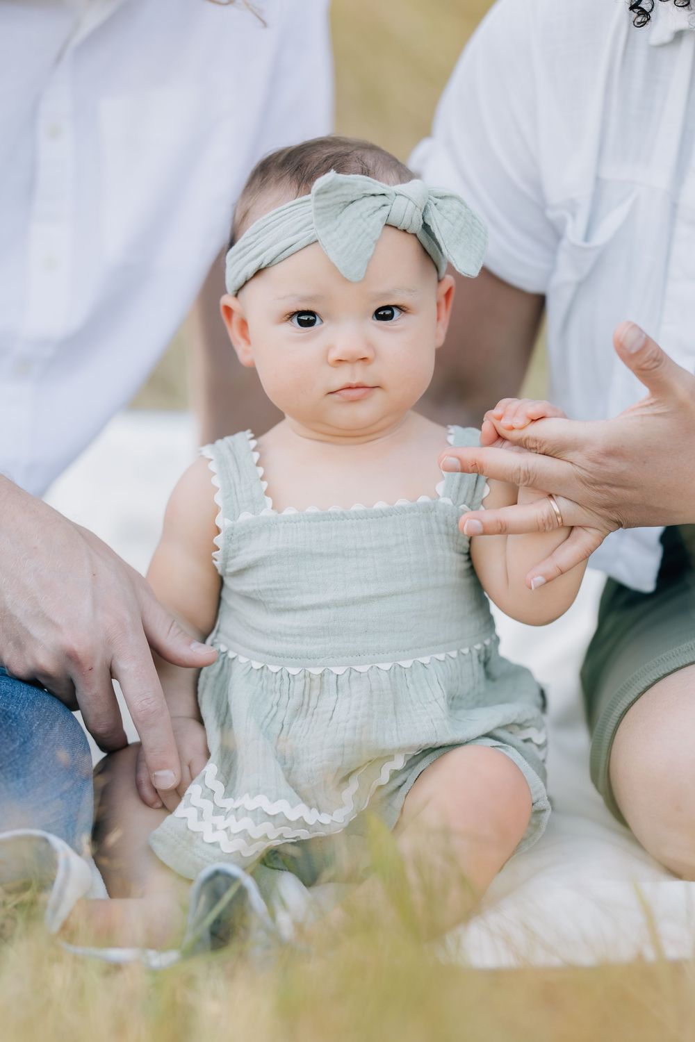 Adorable baby girl in sage green romper and bow headband sits outdoors with parents during a family photo session.