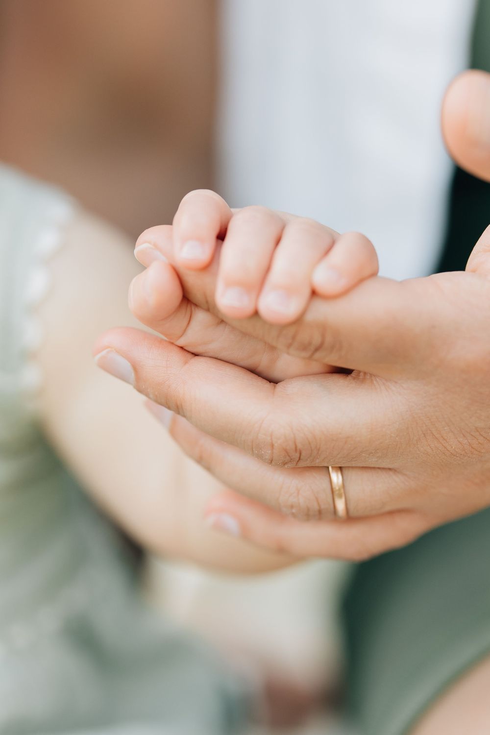 A parent's hand with a gold wedding ring gently holds a tiny newborn baby's hand in a tender moment.
