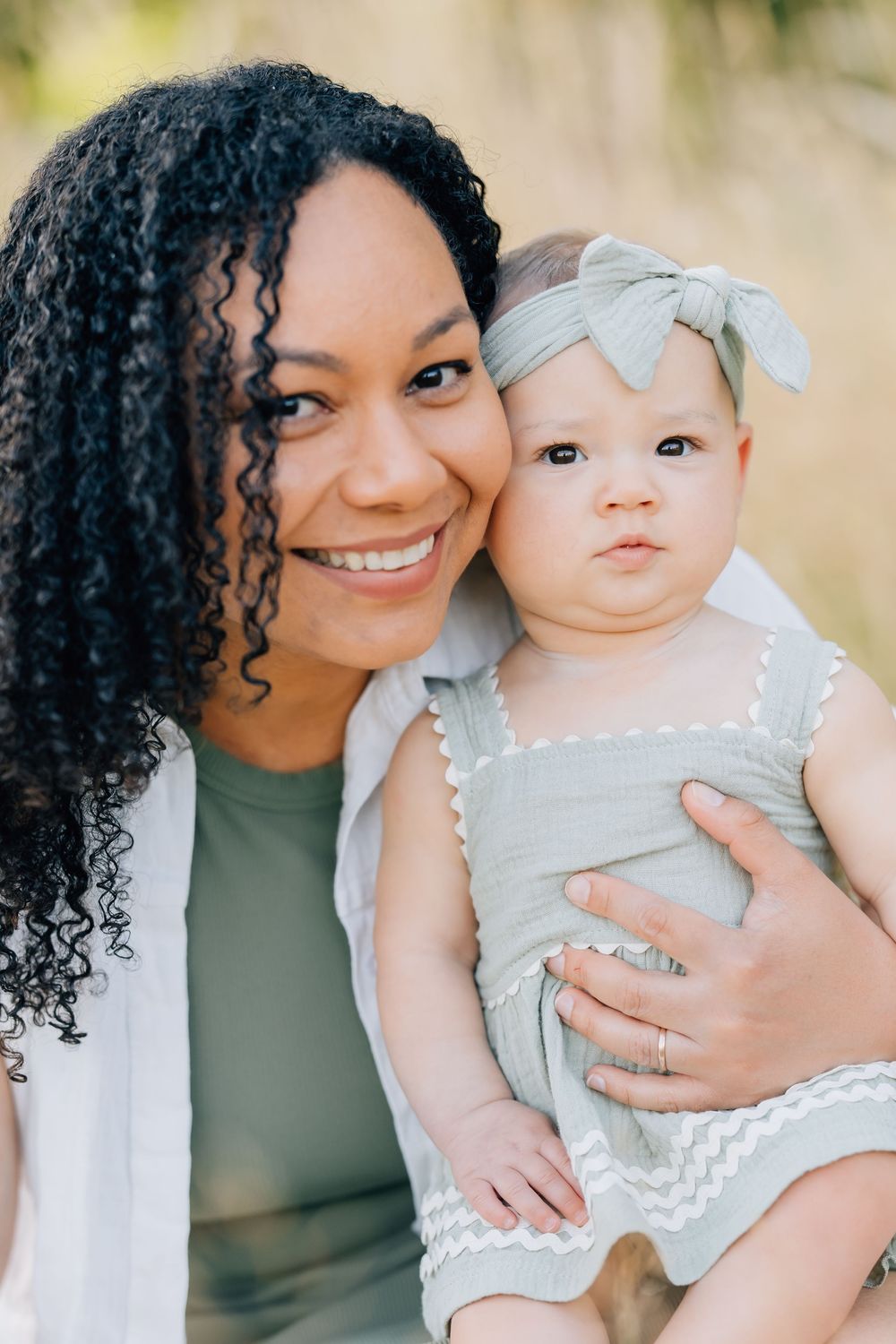 Smiling woman with curly hair holds a cute baby girl in a gray bow headband and dress outdoors.