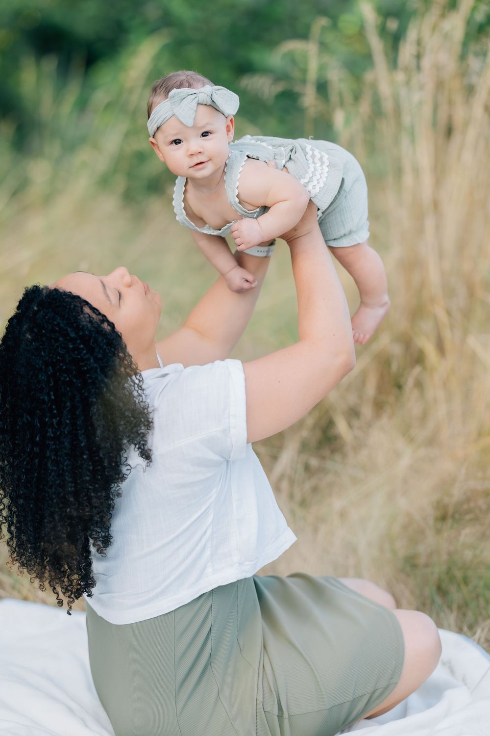 Mother with curly hair lifts smiling baby outdoors in a sunny field, both wearing matching gray and white outfits.