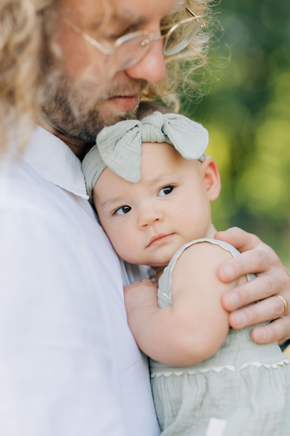 A bearded father with curly hair gently holds his baby girl wearing a sage green bow headband outdoors.