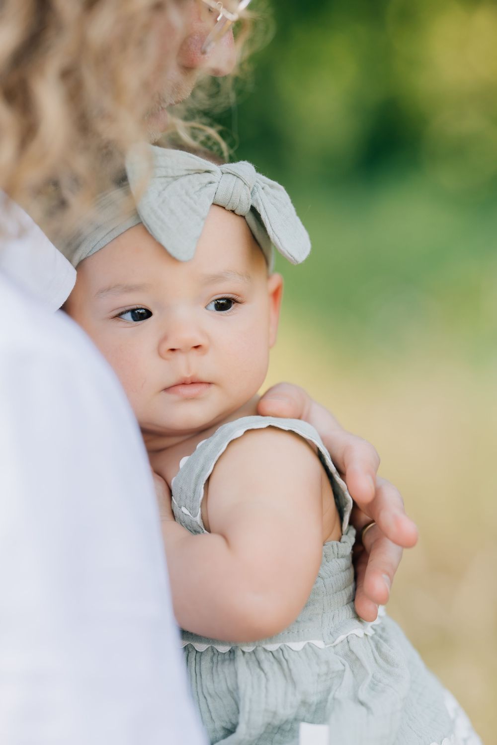 A cute baby girl in a sage green romper and bow headband being held by her curly-haired mother outdoors.
