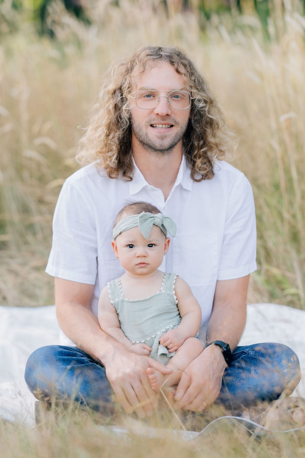 Smiling dad with curly hair sits in golden grass field with his baby daughter wearing a bow headband.