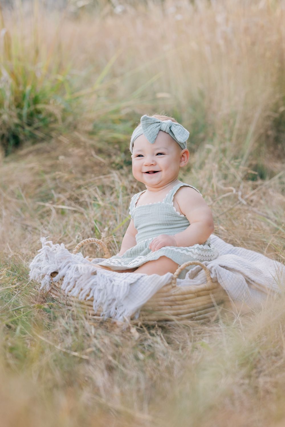 Adorable smiling baby girl in green romper and bow headband sits in a wicker basket in a sunny field.