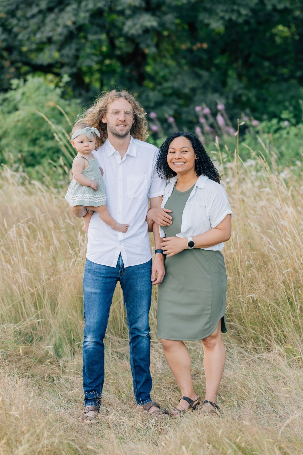 A happy interracial family posing outdoors in a field, man holding baby girl, woman in green dress smiling.