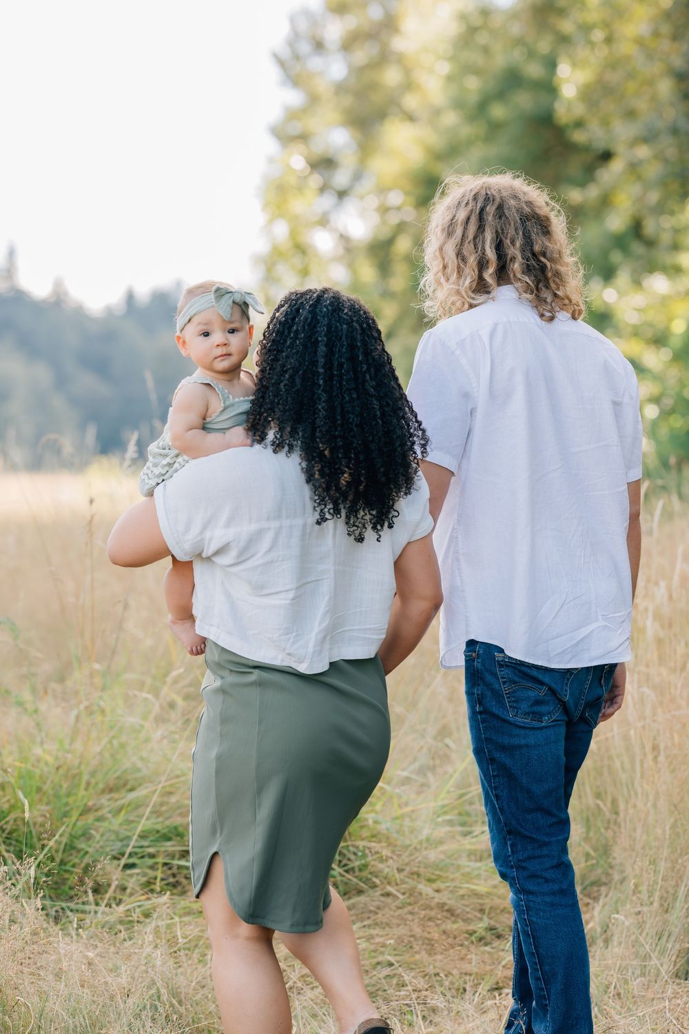 A family of three walks through a sunny meadow, mother carrying baby girl, father beside her, trees in background.