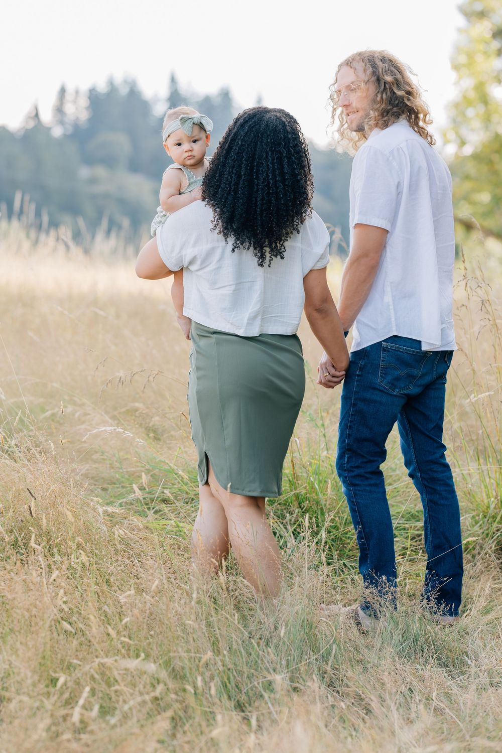 Family of three walks through golden grass field, mother holding baby while father holds her hand.
