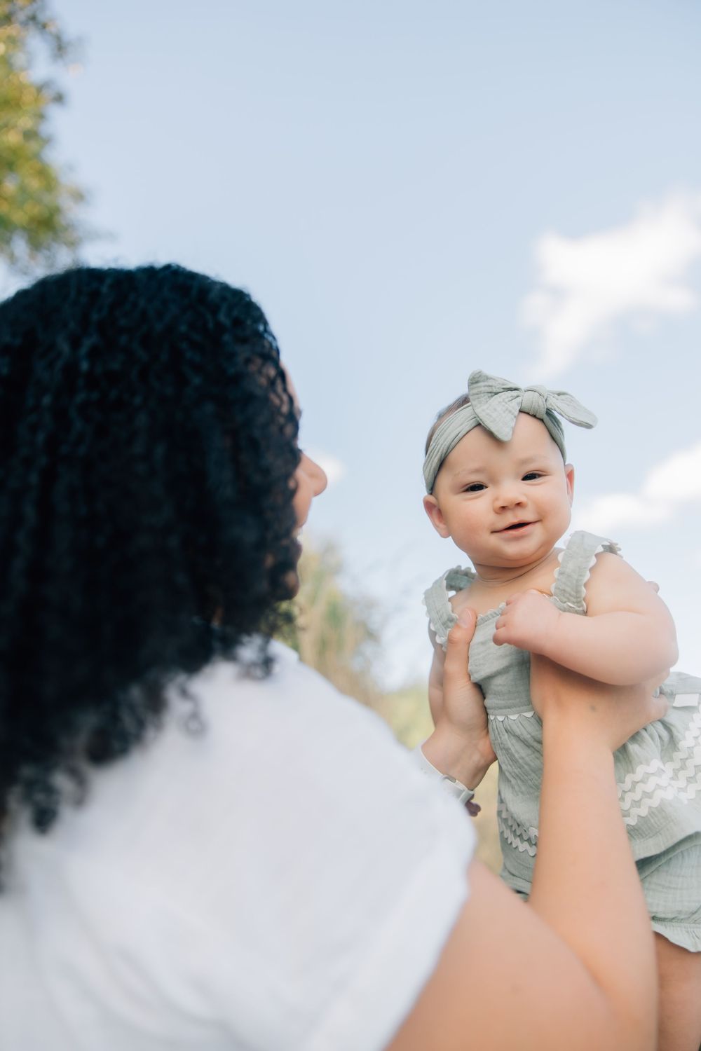 A mother lifts her smiling baby girl wearing a bow headband outdoors under a blue sky.
