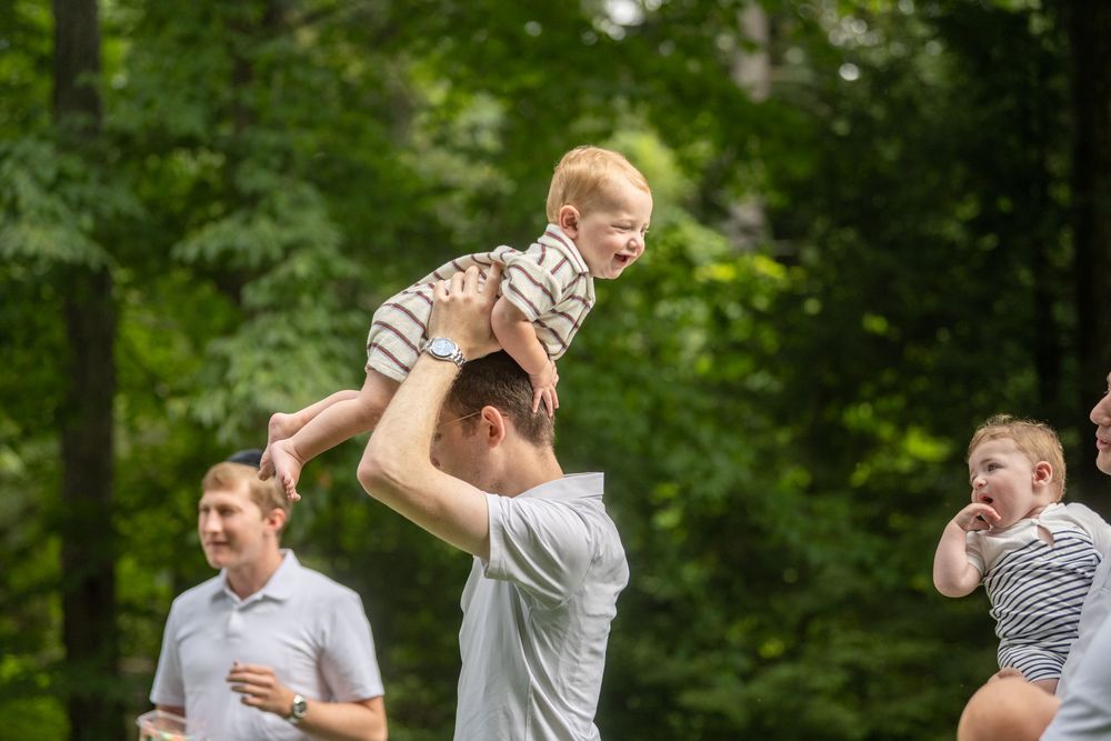 Man lifting happy red-haired toddler overhead outdoors, with smiling adults nearby in a lush green forest setting.