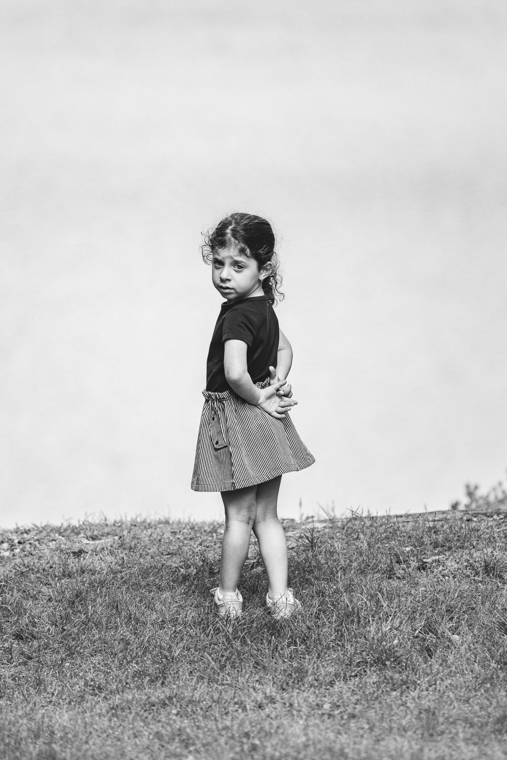 Black and white photo of a young girl in a skirt and t-shirt standing on grass, looking back over her shoulder.