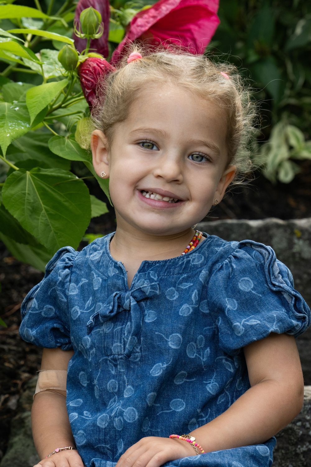 Smiling toddler girl in blue denim dress with bracelets sitting outdoors near pink flowers and green plants.