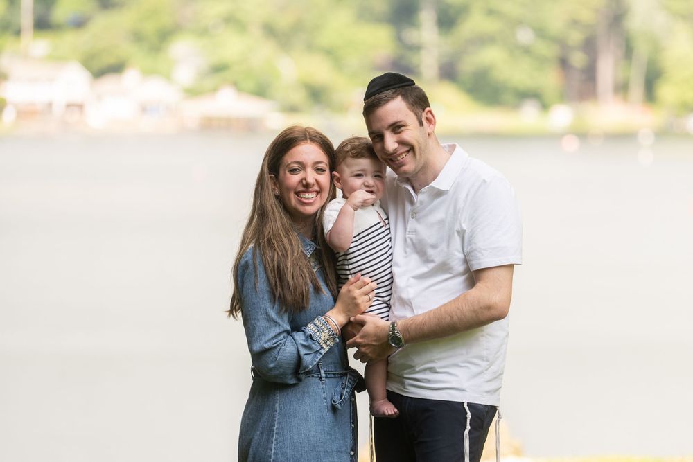 Happy couple smiling outdoors near a lake, holding their baby in a striped outfit on a bright day.