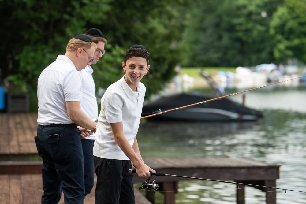 Two teenage boys fishing together on a wooden dock by a lake surrounded by green trees.