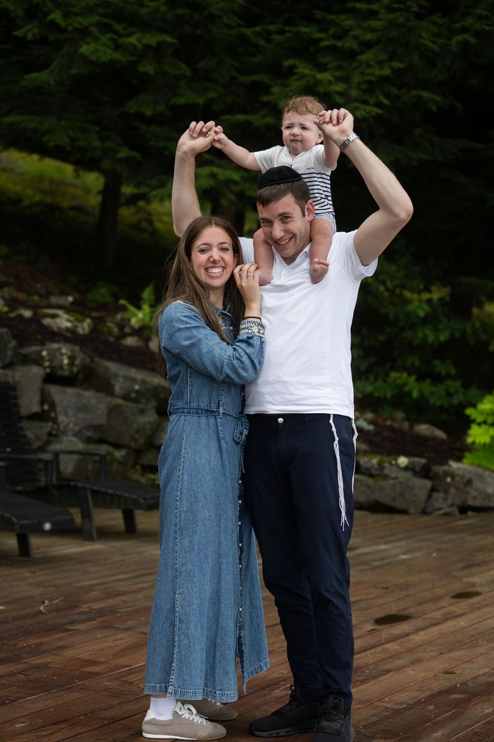Happy young family with toddler on dad's shoulders posing outdoors on wooden deck surrounded by lush greenery.