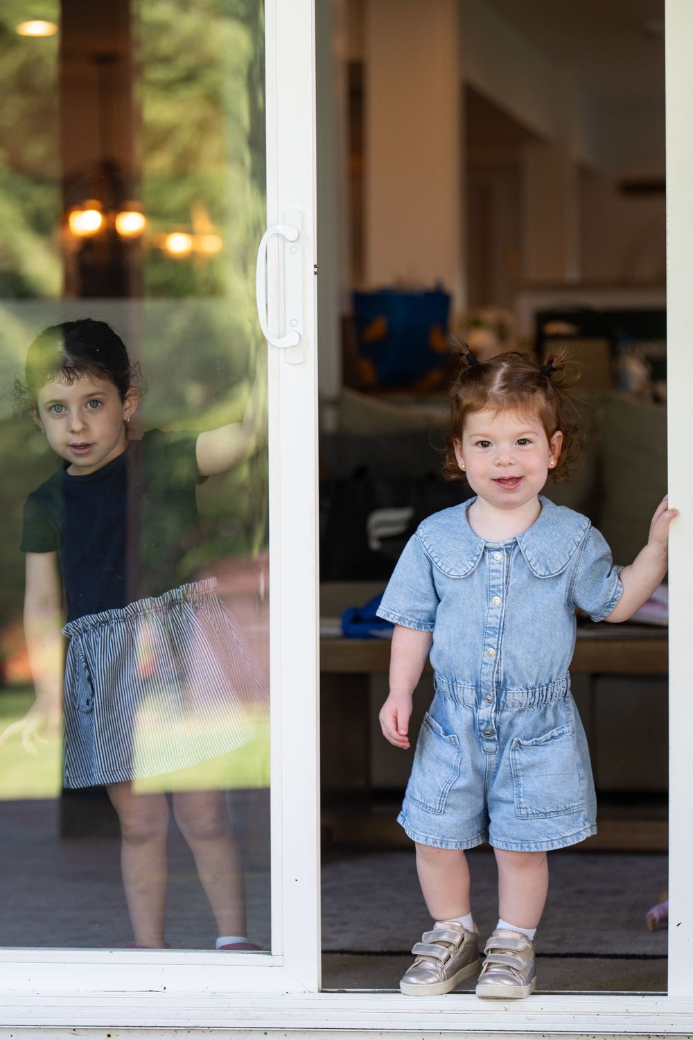 A toddler girl in denim romper stands at sliding glass door while a boy peers through the window outside.