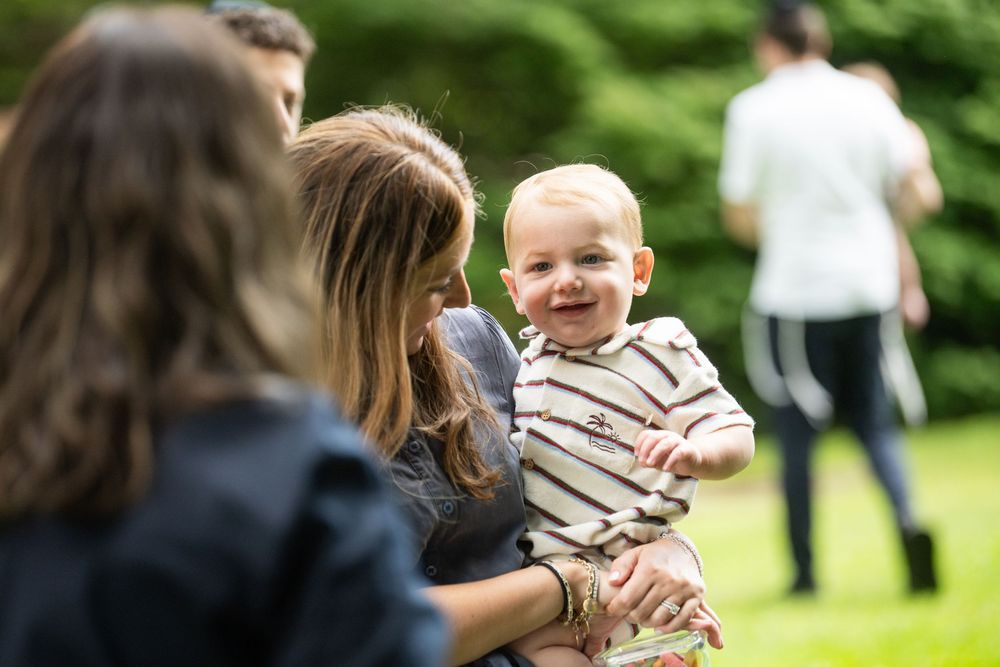 A smiling baby being held by a woman outdoors on a green lawn with people in the background.