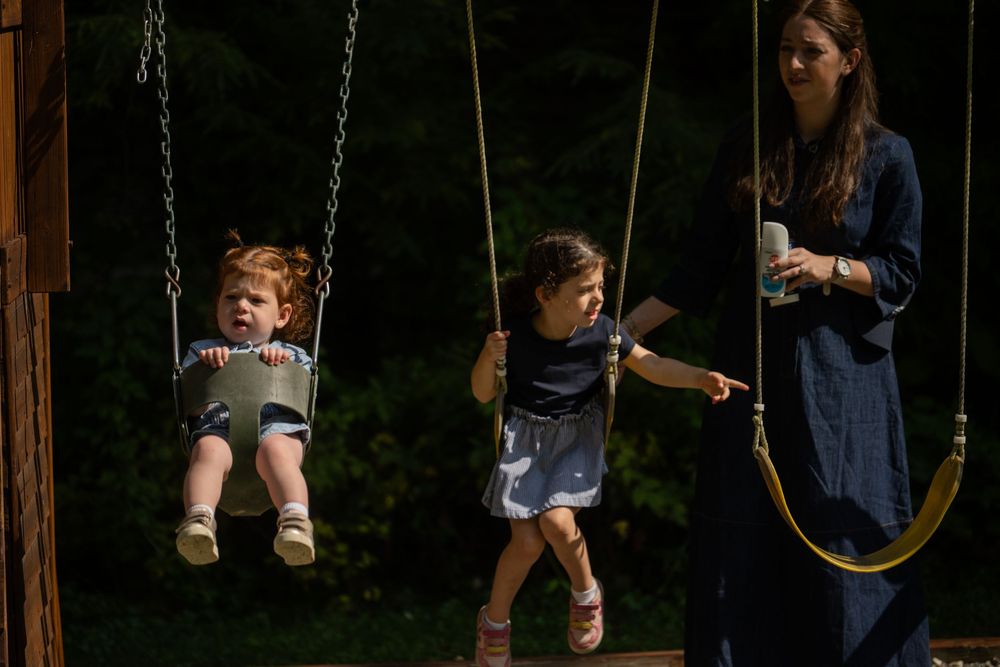 Woman pushes two young girls on outdoor swings at a playground surrounded by lush green trees.