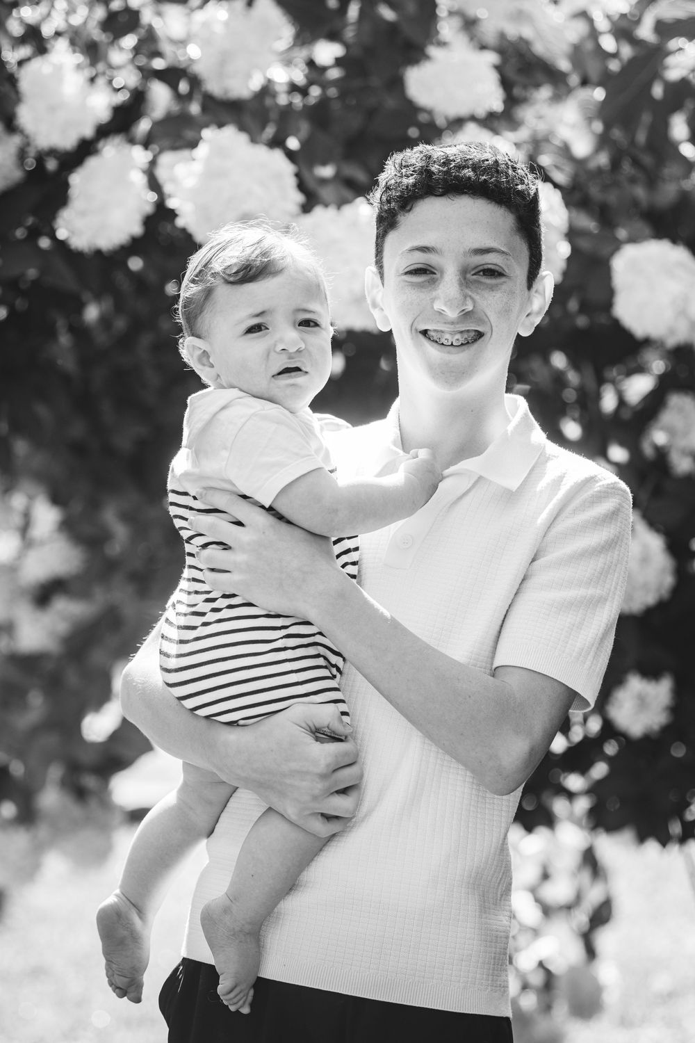 Black and white photo of a smiling teen with braces holding a baby outdoors near blooming flowers.