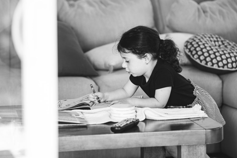 Black and white photo of a young girl sitting at a wooden coffee table, focused on drawing or writing in a book.