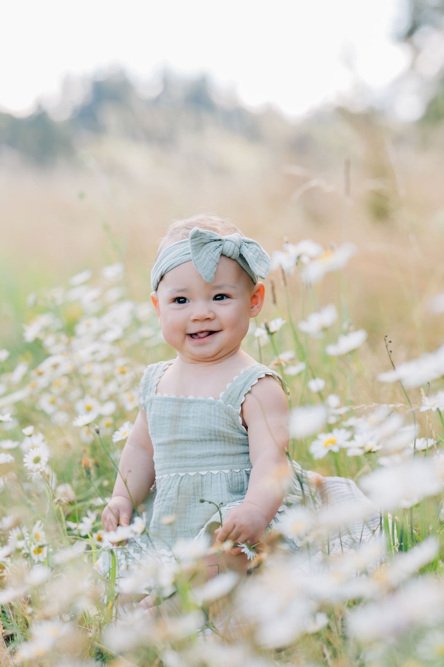 Smiling baby girl in sage green romper and bow headband sitting among white wildflowers in a sunny meadow.