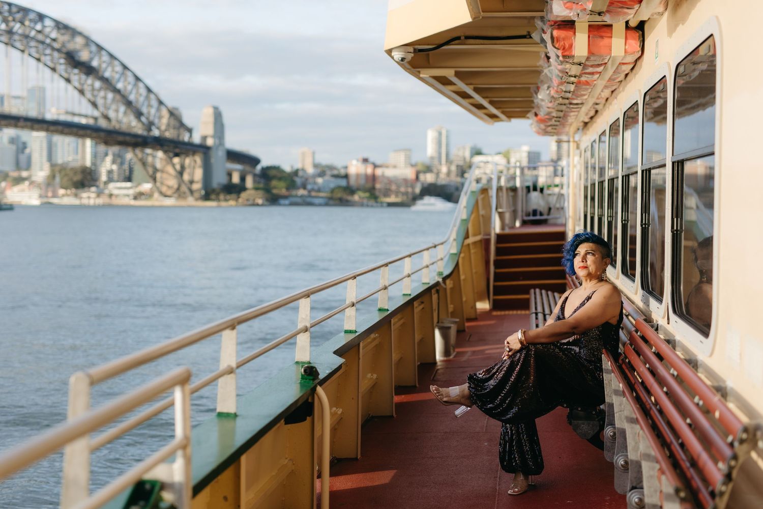 A woman in elegant attire sits on a ferry deck with Sydney Harbour Bridge and city skyline in the background.