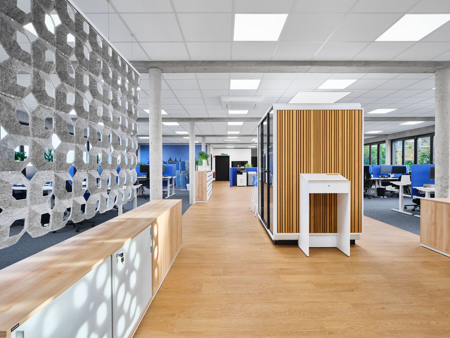 Modern open-plan office interior with wood flooring, a honeycomb acoustic wall, a wooden meeting pod and workstations in the background.