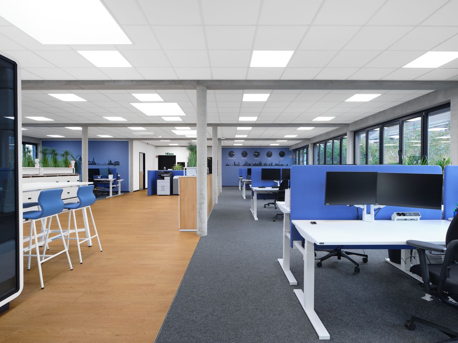 Modern open-plan office with blue partitions, white desks, dual monitors, high stools and wood flooring under bright ceiling lights.