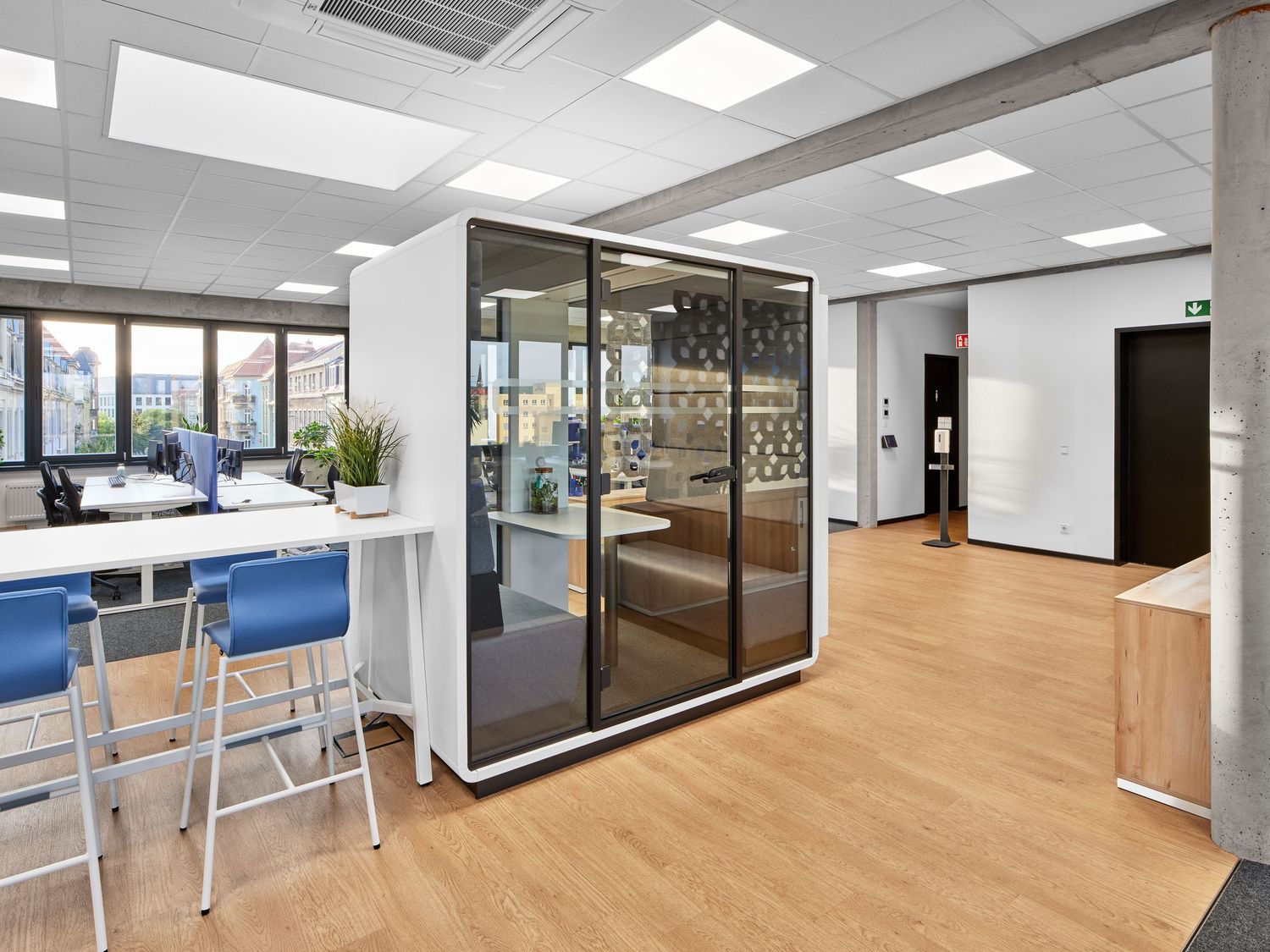 Modern open office with a glass meeting pod, blue bar stools at a white standing table, wood flooring and bright ceiling lights.