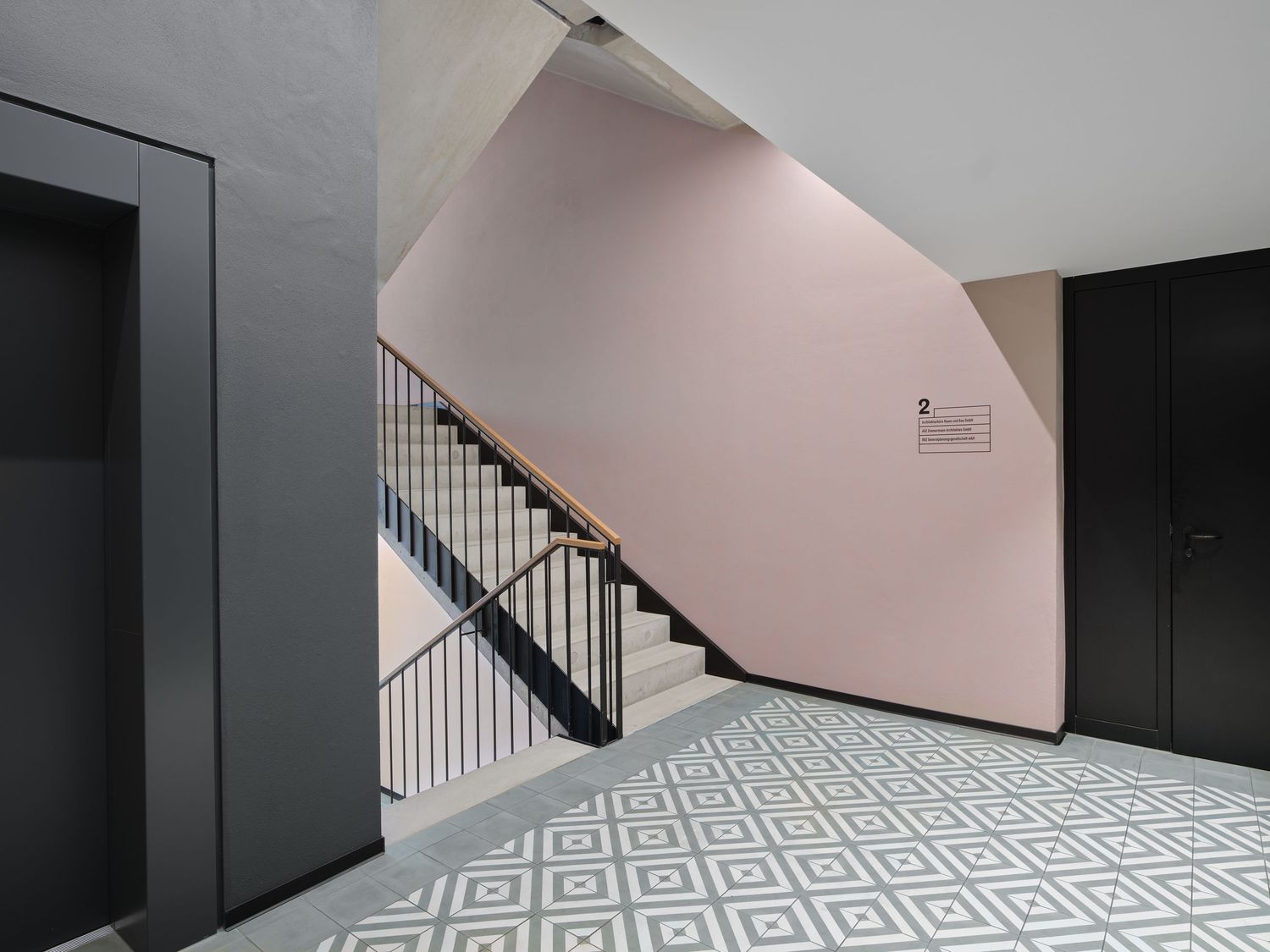 Modern stairwell with pink walls, geometric black and white tiled floor, dark metal railing and charcoal doors.