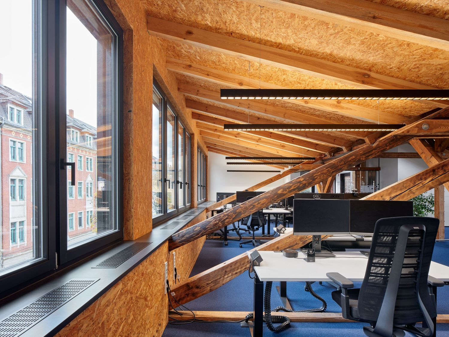 Modern open-plan office with exposed timber beams, wood-paneled ceiling, large windows and ergonomic chairs at white desks.