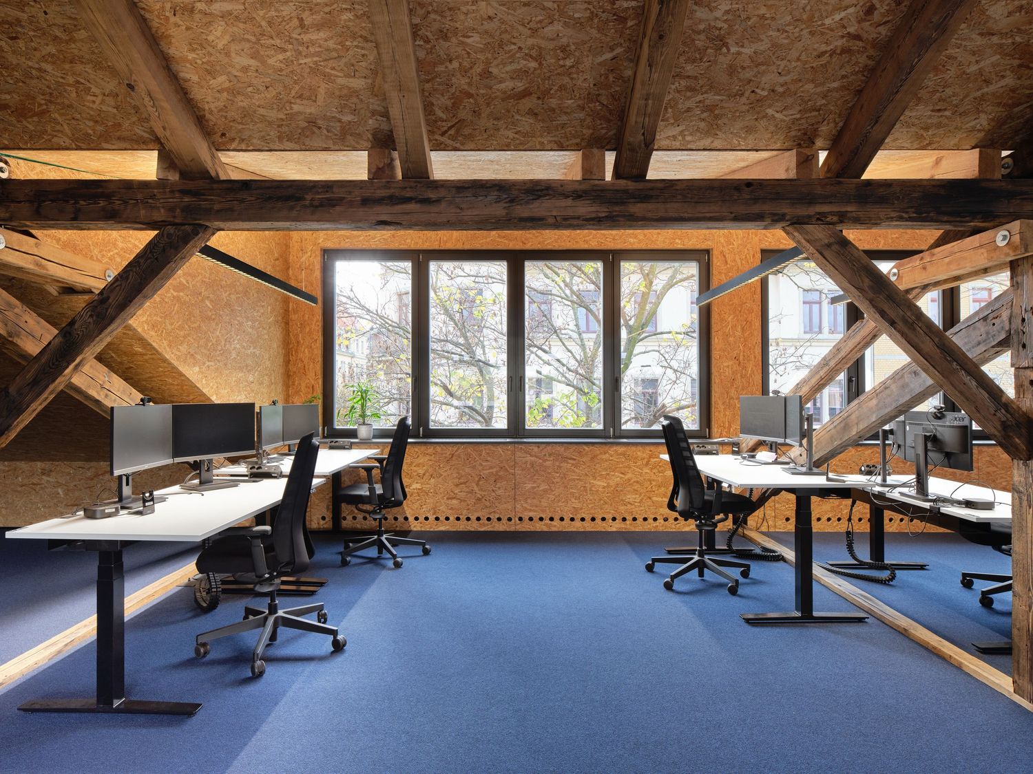 Spacious loft office with rustic wooden beams, brick walls, blue carpet and rows of desks with monitors beside tall windows.