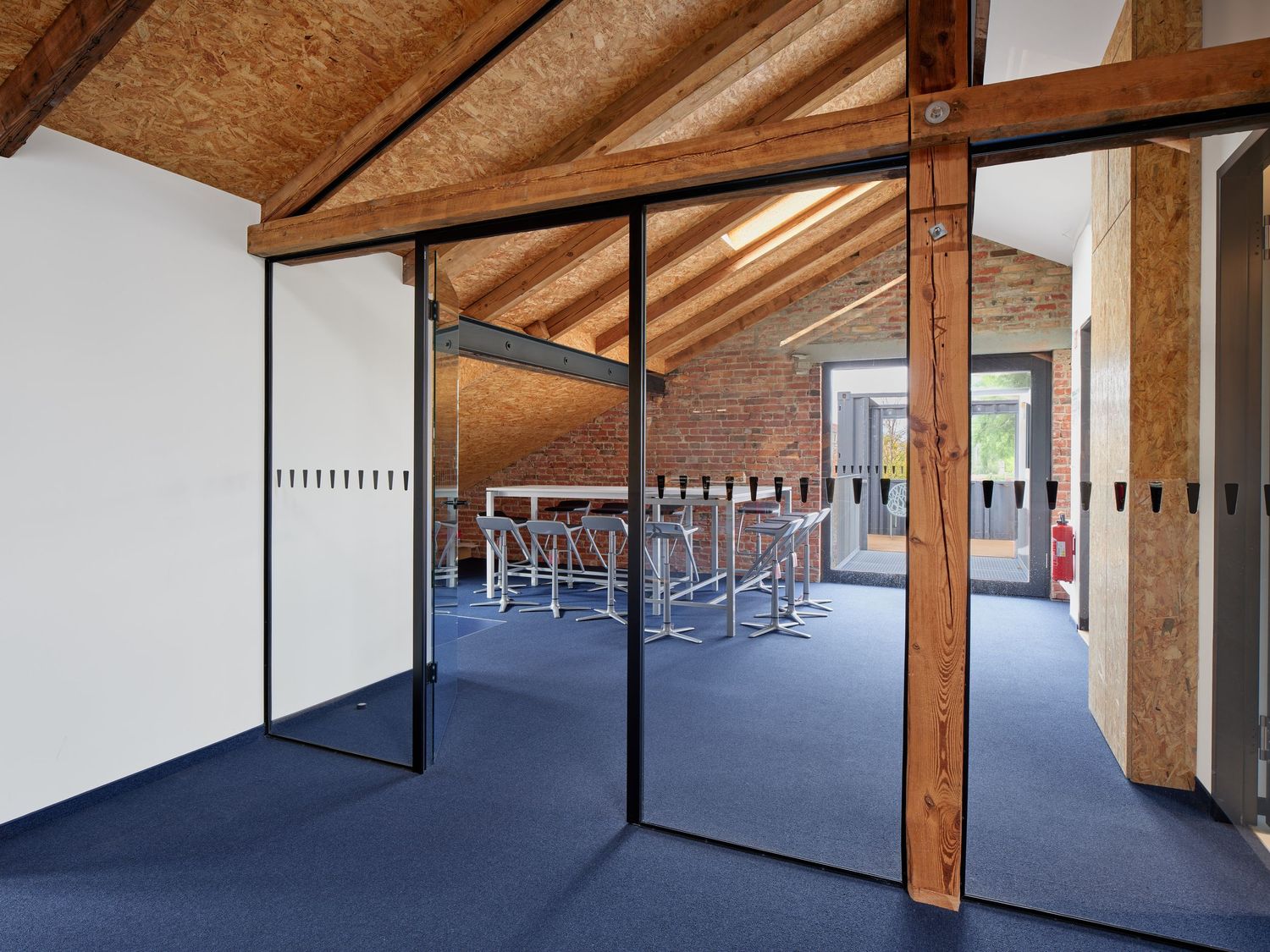 Modern attic meeting room with glass partition walls, exposed timber beams, brick wall, blue carpet and a high table with stools.