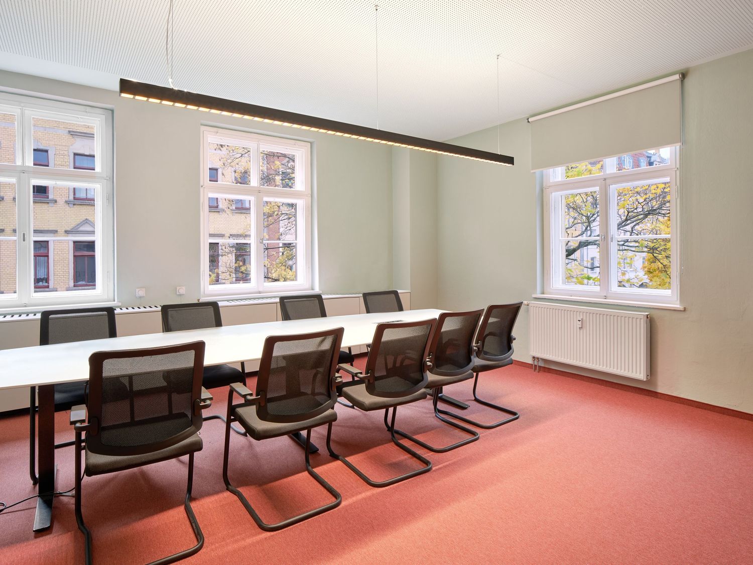Bright meeting room with a long white table, black mesh chairs, red carpet, large windows and a suspended linear light fixture.