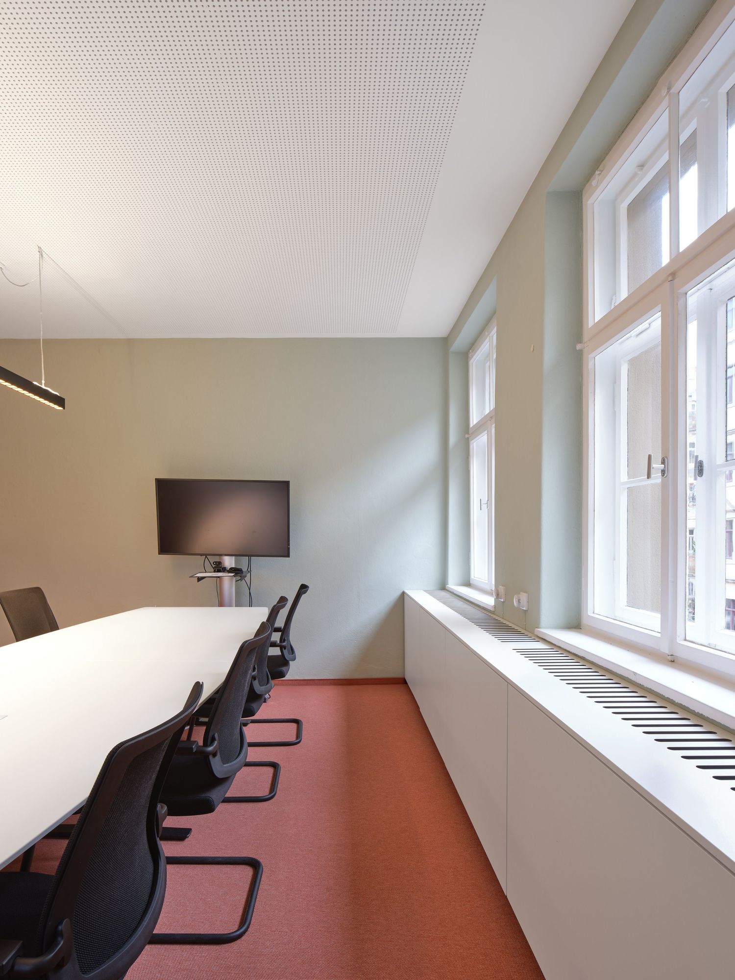 Modern conference room with a long white table, black mesh chairs, wall-mounted monitor, orange carpet and tall windows with natural light.