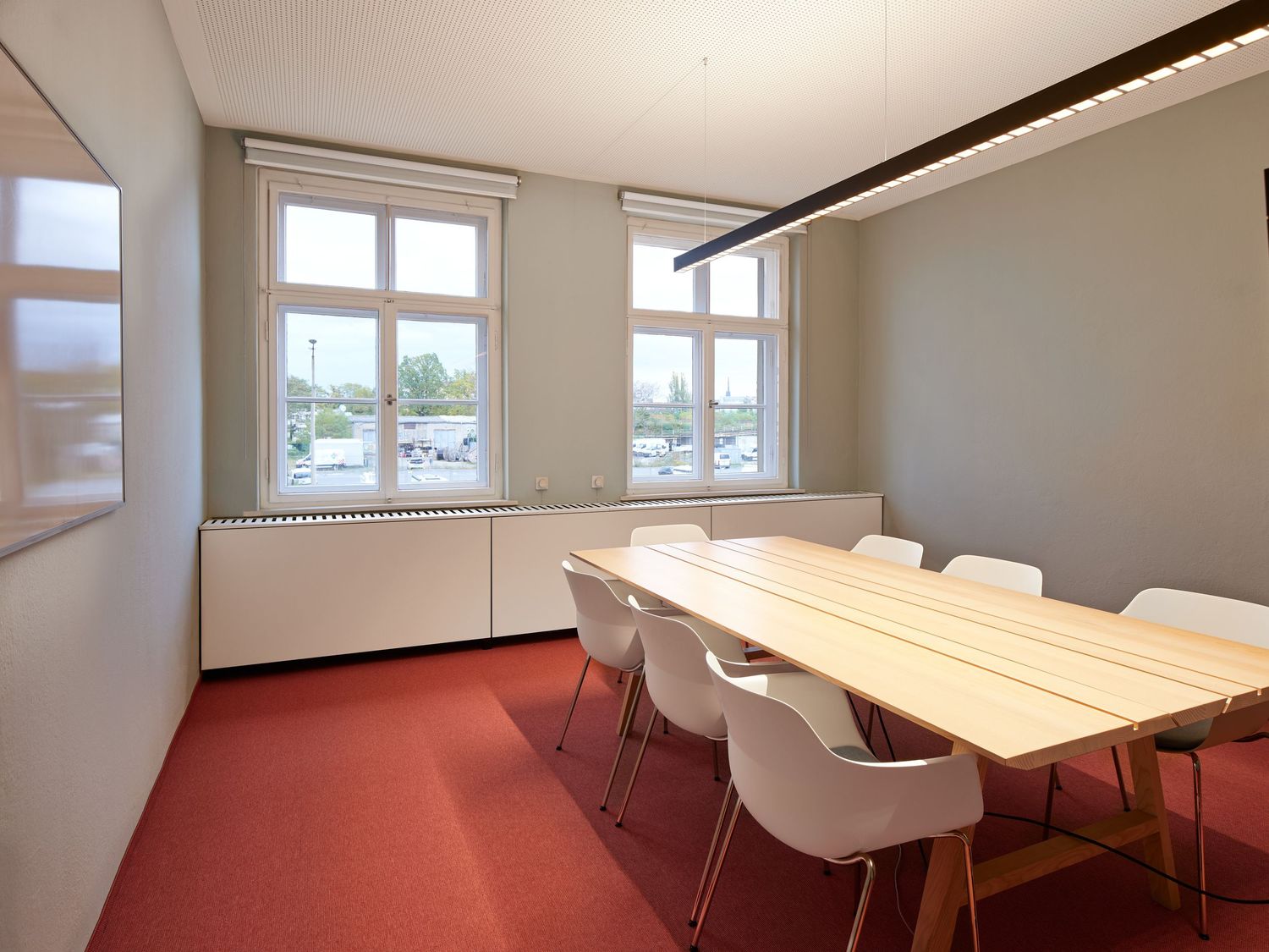 Bright modern meeting room with a long light wood table, white chairs, red carpet and large windows overlooking a parking area.