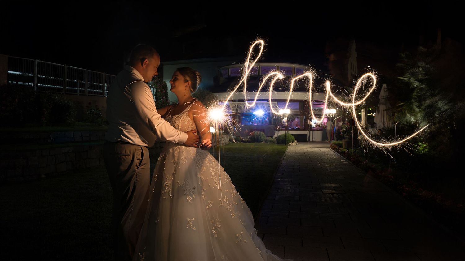 Bride and groom embrace at night while a sparkler light trail spells the word love in the air beside a garden path.