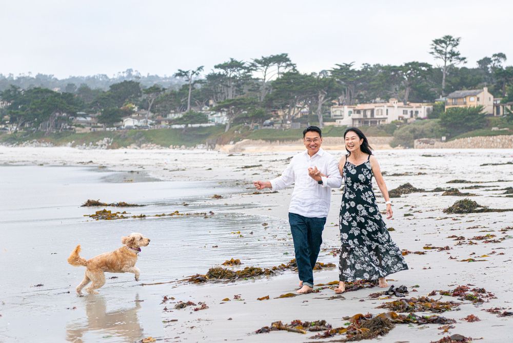 A smiling couple walks barefoot along a seaweed-strewn beach while a golden retriever runs beside them on an overcast day.